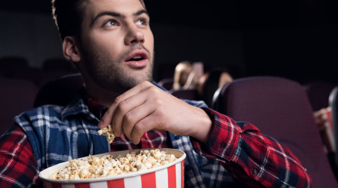 Man sitting in a movie theater holding a striped bucket of popcorn, looking surprised or captivated