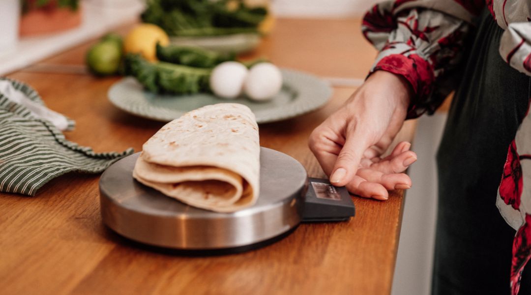 Close-up of a tortilla being weighed on a kitchen scale, with fresh vegetables and eggs in the background