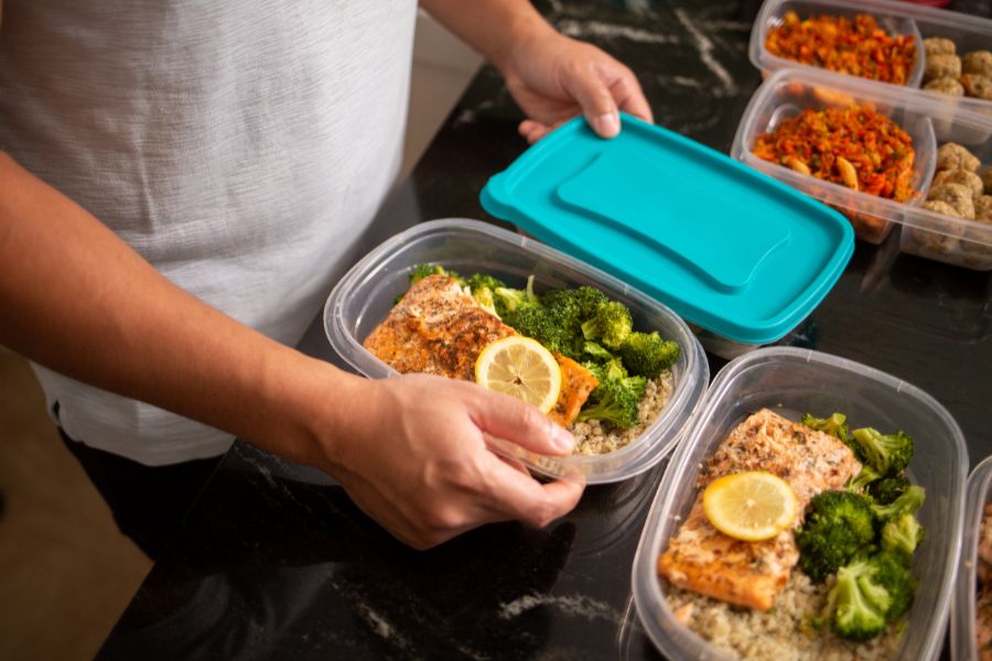 Person placing a lid on a meal prep container with salmon, broccoli, and quinoa