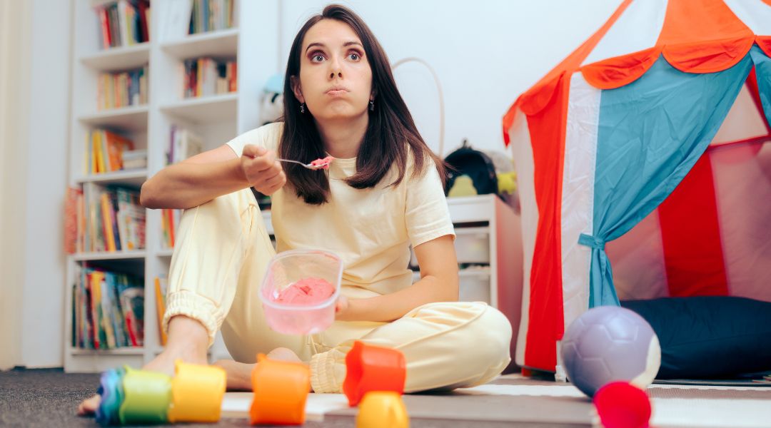 Woman sitting on the floor of a playroom, eating ice cream from a container with toys scattered around