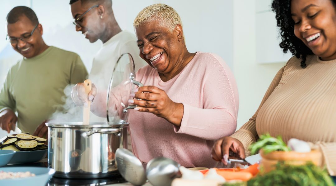 Group of four people laughing and cooking together in a kitchen, preparing a meal with fresh vegetables