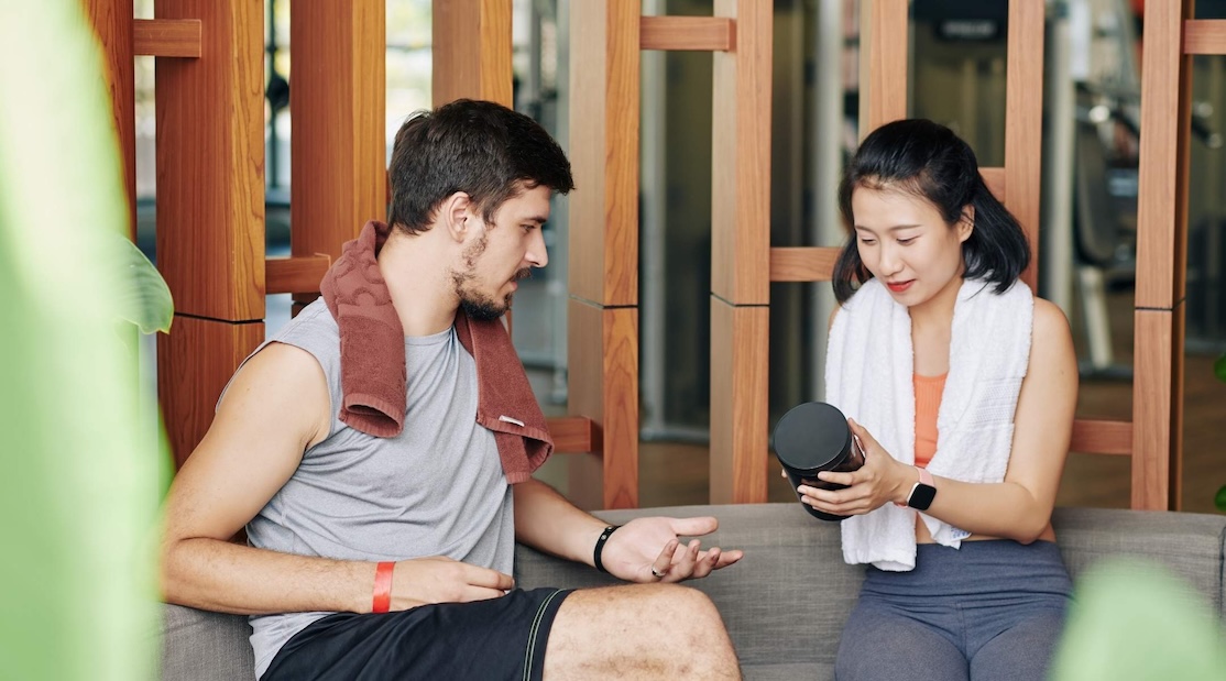 A man and a woman in workout clothes sitting on a bench, discussing a supplement container she is holding, in a gym setting.