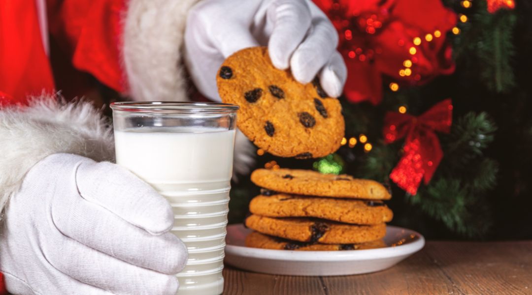 Santa's gloved hand holding a cookie above a glass of milk, with a stack of cookies and Christmas decorations in the background