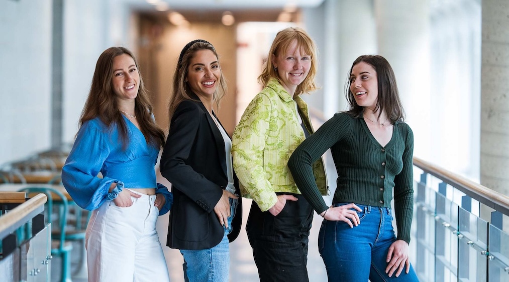 Four women smiling and standing in a hallway with their hands on their hips, wearing casual and stylish outfits.