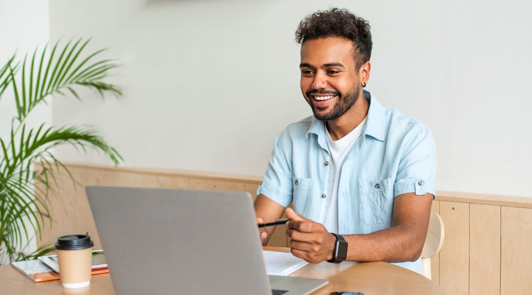 Man smiling at his laptop while working, with a coffee cup and notepad on the desk in a cozy setting