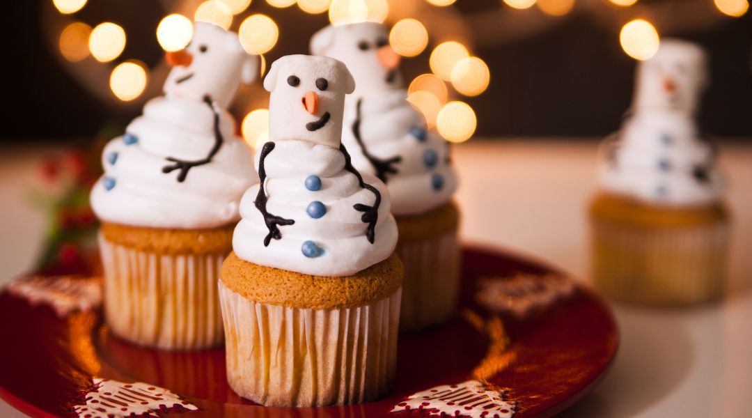 Festive cupcakes decorated with marshmallow snowmen, featuring smiling faces and twig-like arms, on a red plate