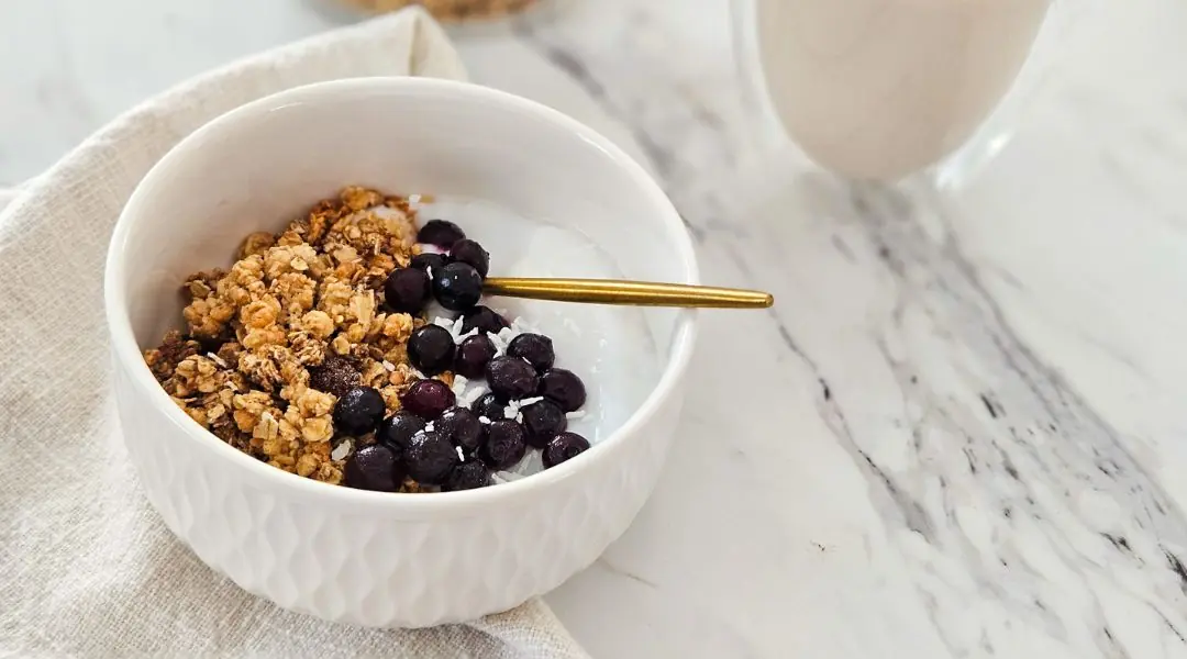 Bowl of yogurt topped with crunchy granola, blueberries, and coconut, with a gold spoon