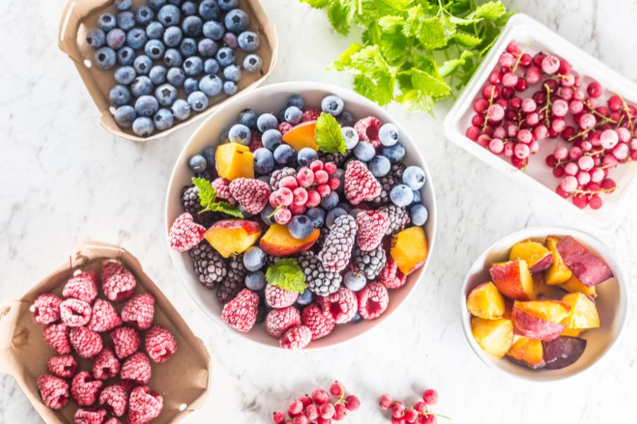 Assorted frozen berries and sliced fruits in bowls on a white background