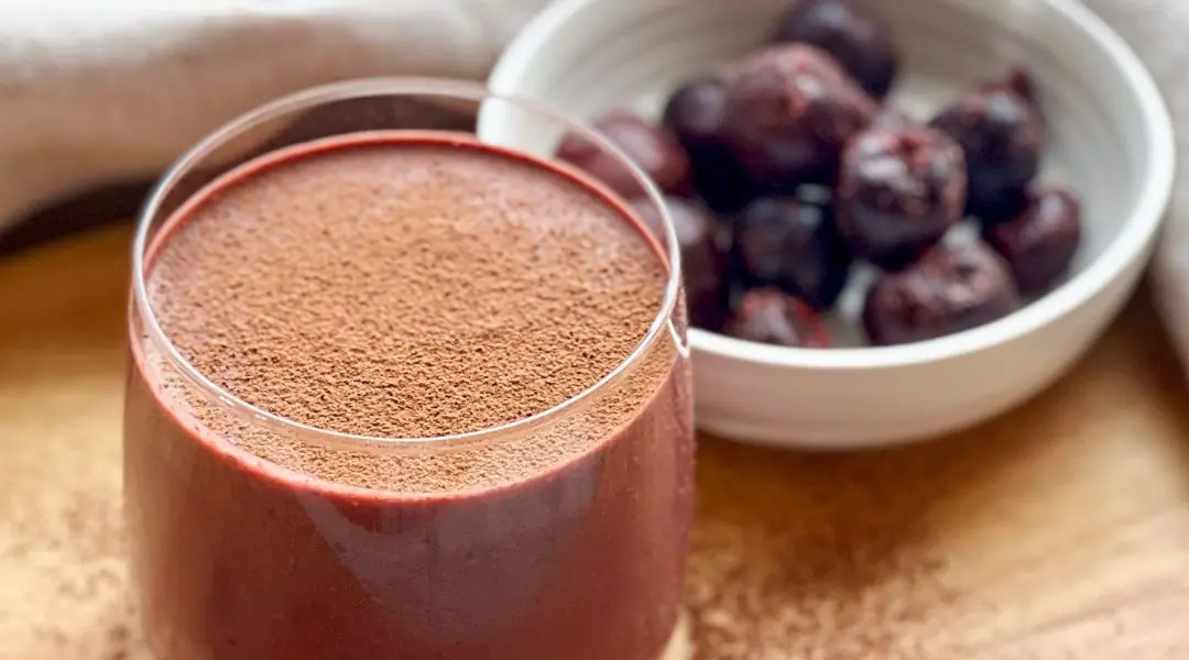 Thick cherry and cocoa smoothie in a glass, with frozen cherries in the background
