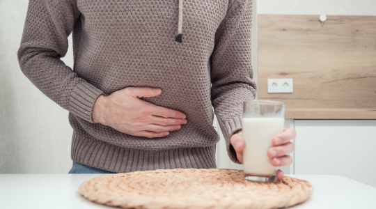 A person holding their stomach with one hand, expressing discomfort, while holding a glass of milk with the other hand, resting on a table.