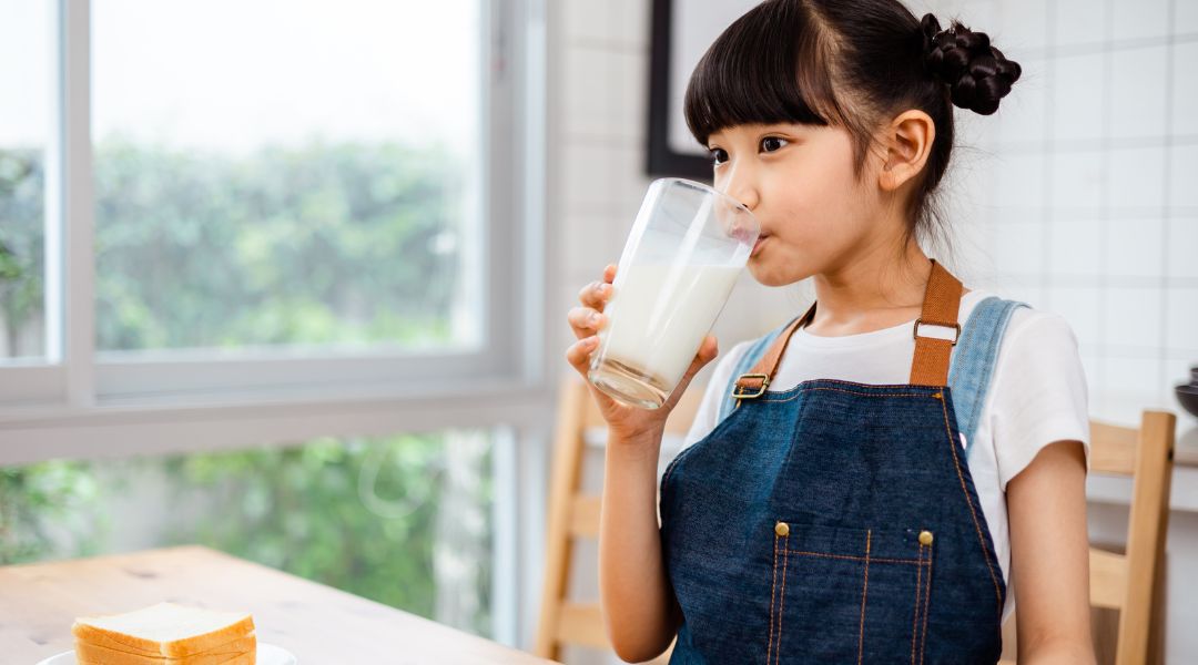 A child in a blue apron drinks milk from a glass, with a plate of bread on a table near a bright window and greenery outside.