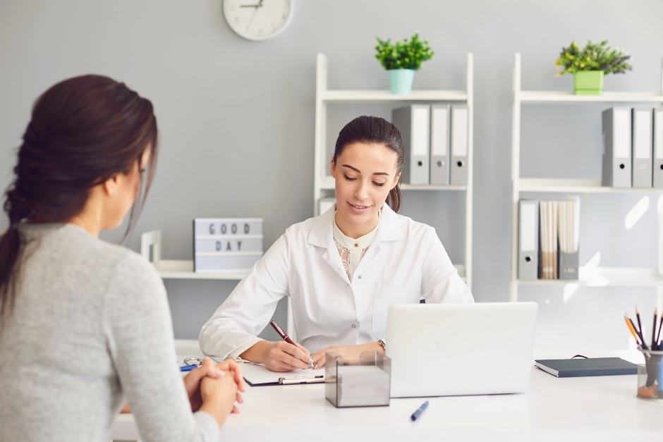 Une nutritionniste-diététiste en consultation avec une patiente à son bureau, prenant des notes en souriant, avec un ordinateur portable et des dossiers en arrière-plan.  A female dietitian taking notes during a consultation with a patient in a modern office.