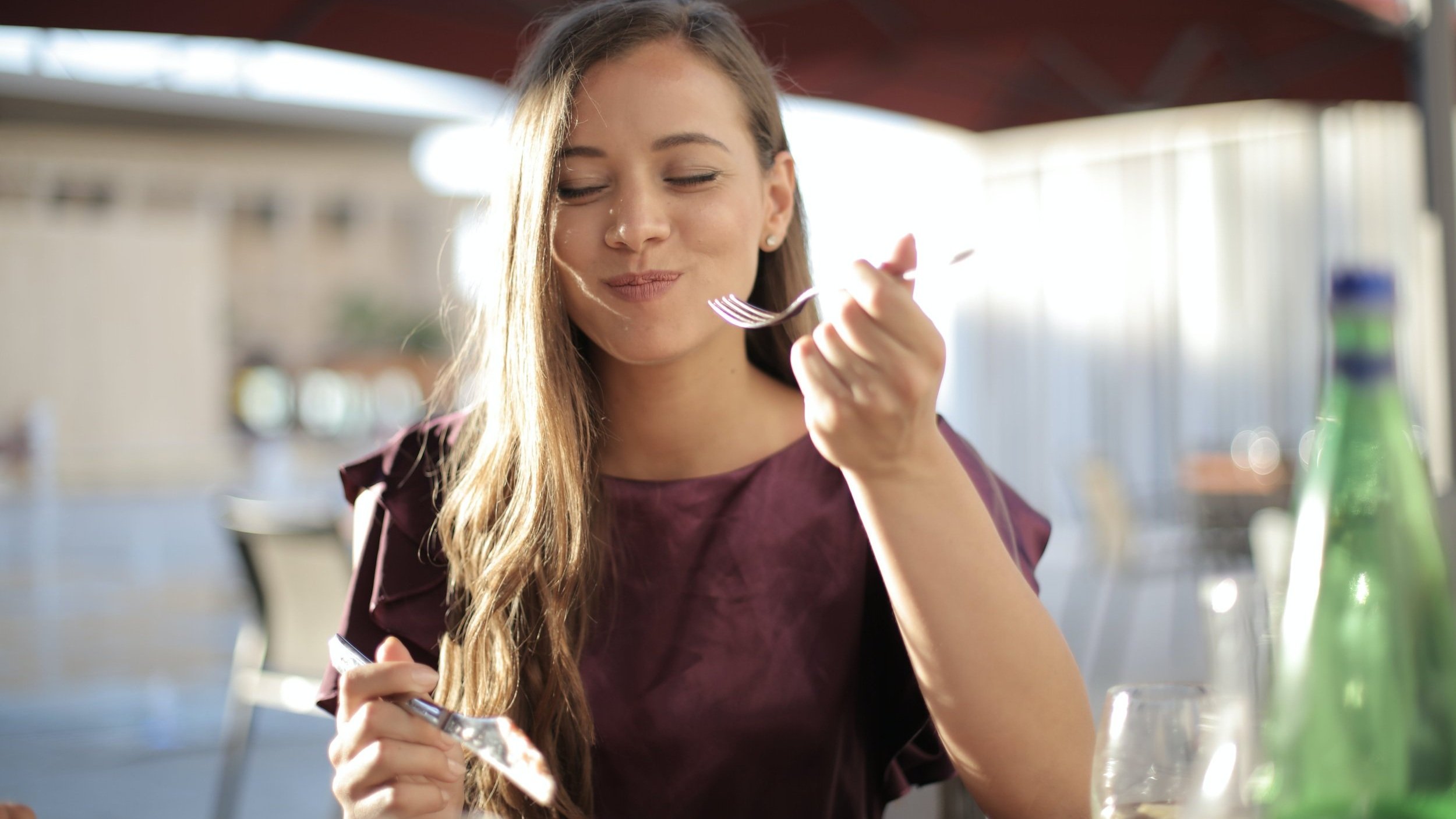 Une femme savourant un repas en extérieur, souriant les yeux fermés, tenant une fourchette dans une main.  A woman enjoying a meal outdoors, smiling with her eyes closed while holding a fork in one hand.