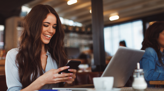 A smiling young woman sitting in a café, using a cellphone while having an open laptop in front of her.