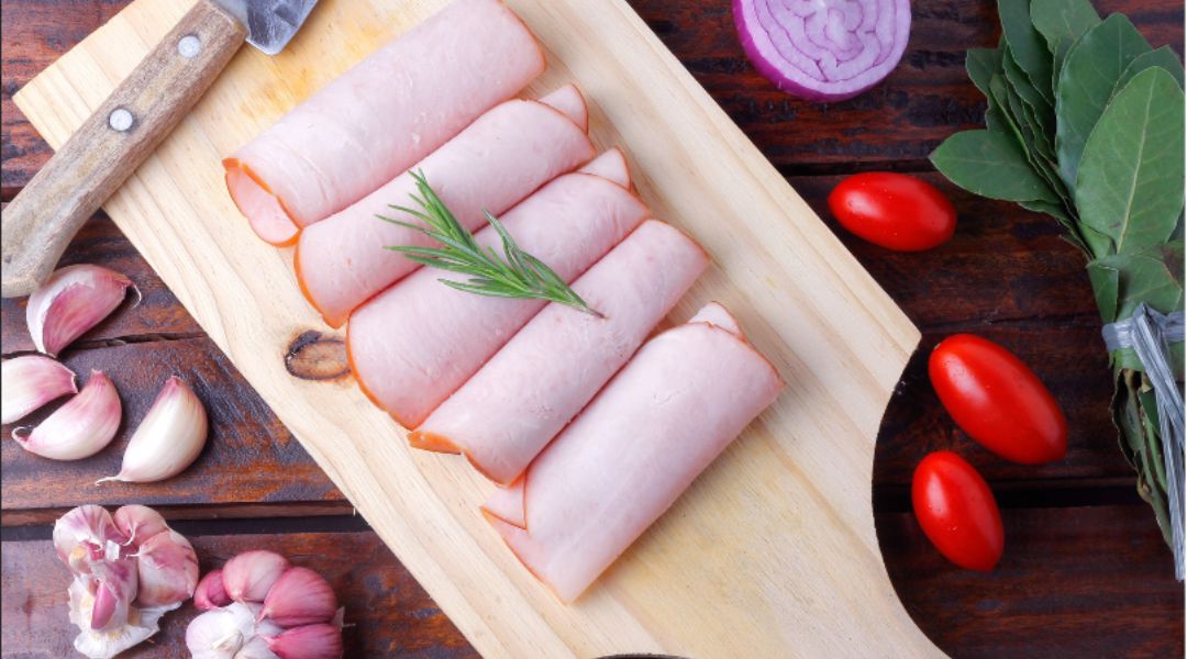 Thinly sliced rolled ham garnished with rosemary on a wooden cutting board, surrounded by garlic, cherry tomatoes, red onion, and bay leaves.