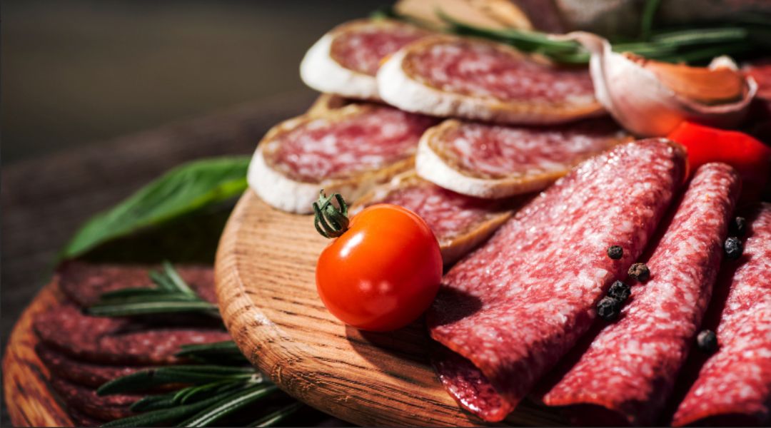 A wooden platter displaying sliced salami, cured meats, and camembert with cherry tomatoes, rosemary sprigs, and peppercorns.