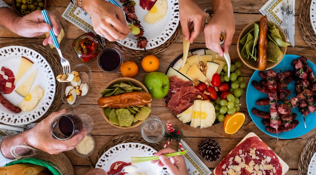 A festive table filled with plates of cheese, cured meats, grilled skewers, fresh fruit, nuts, and dips, surrounded by people serving themselves.