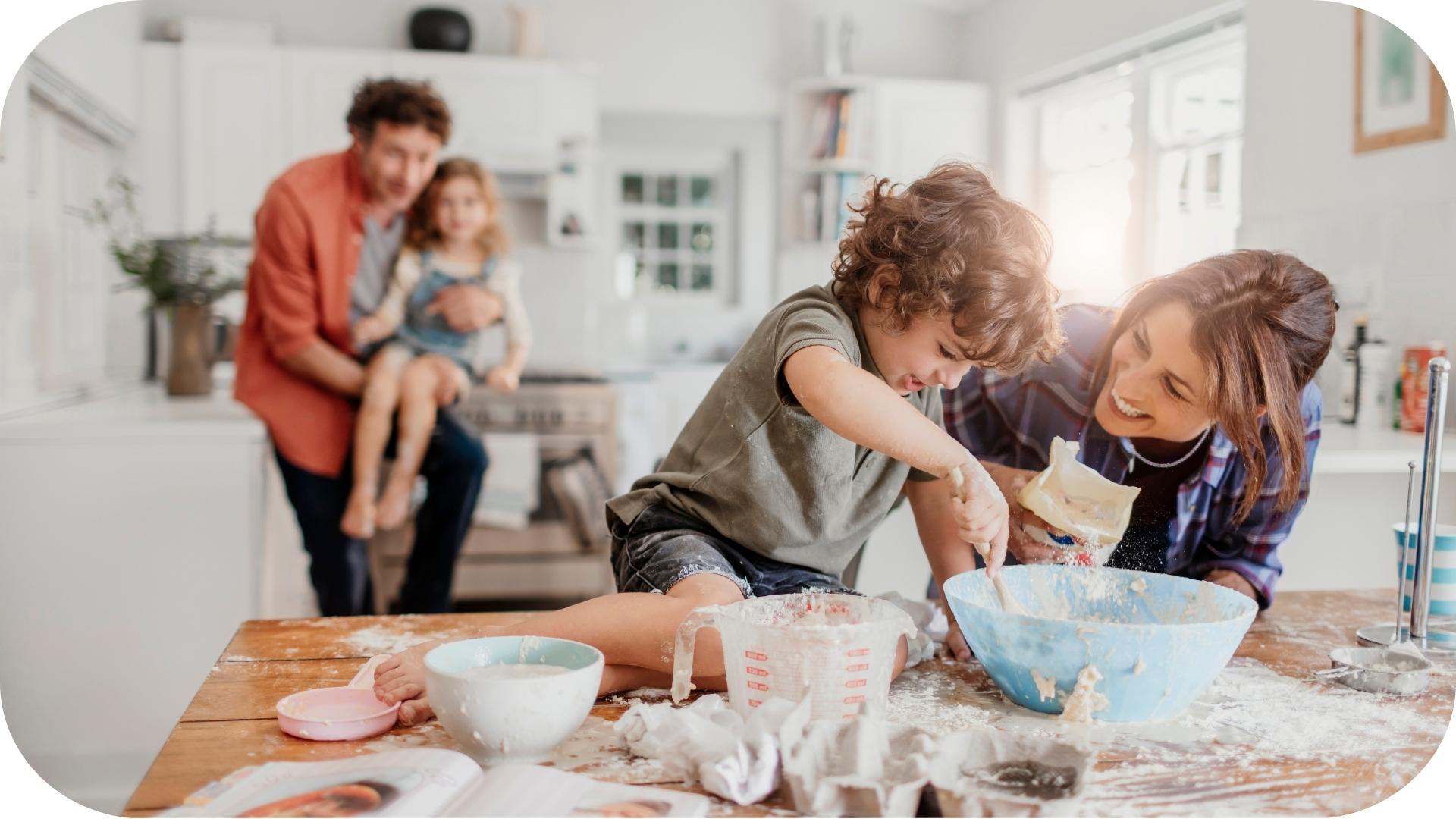 A cheerful family baking together in a bright kitchen. A mother and young boy mix ingredients in a bowl on a messy table, while a father holds a little girl in the background