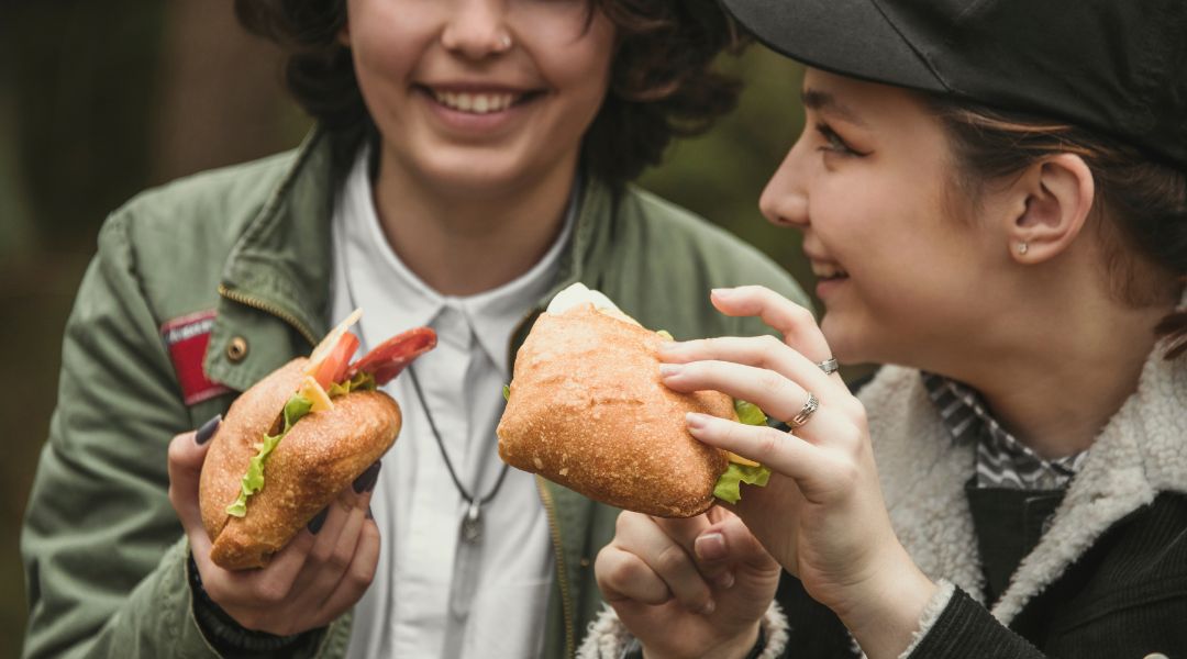 Two young women smiling and holding sandwiches filled with lettuce, tomatoes, and other fresh ingredients, enjoying an outdoor meal.