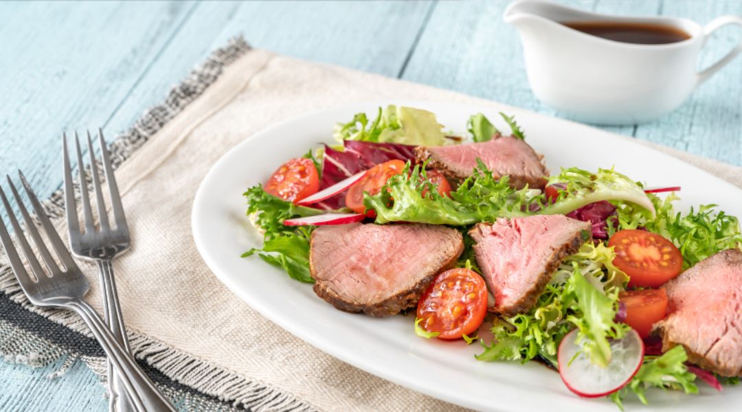 A fresh salad with slices of beef tenderloin, mixed greens, cherry tomatoes, and radishes, served with a side of vinaigrette.