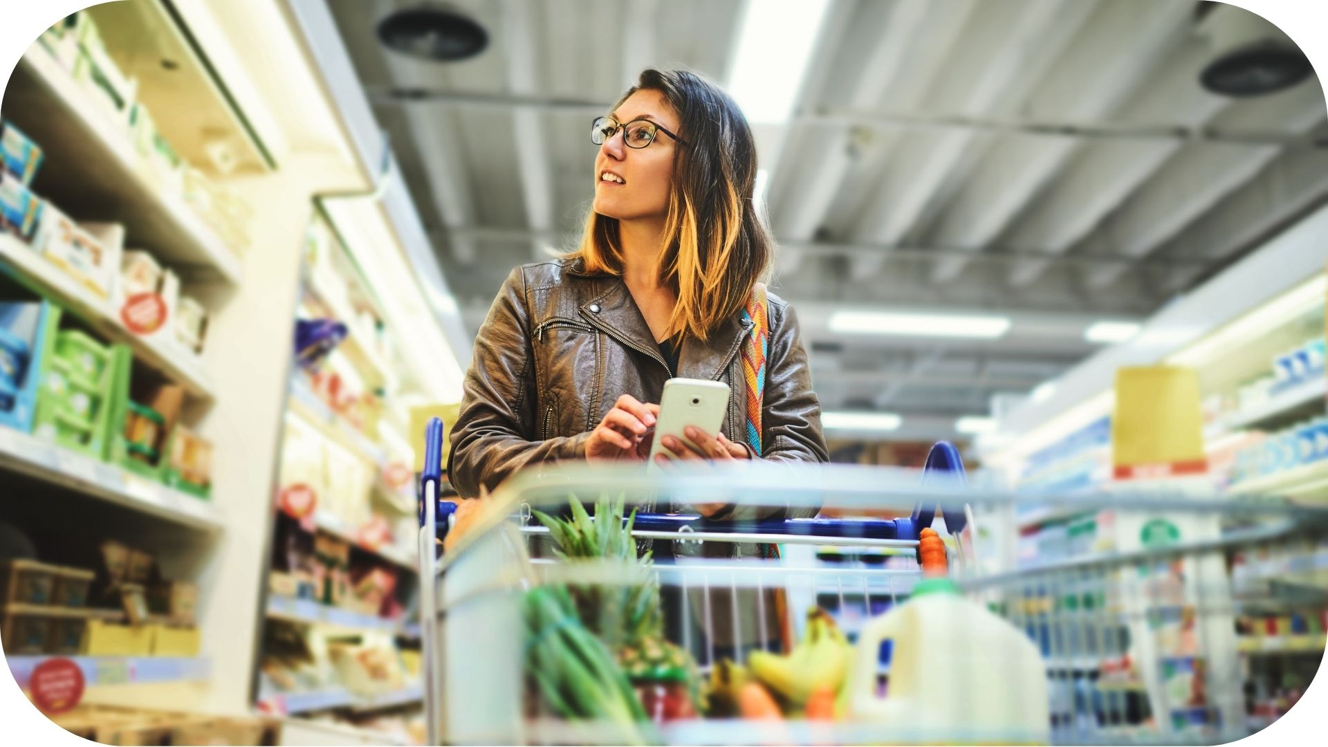 Woman shopping in a supermarket, pushing a cart loaded with fresh produce and holding a smartphone