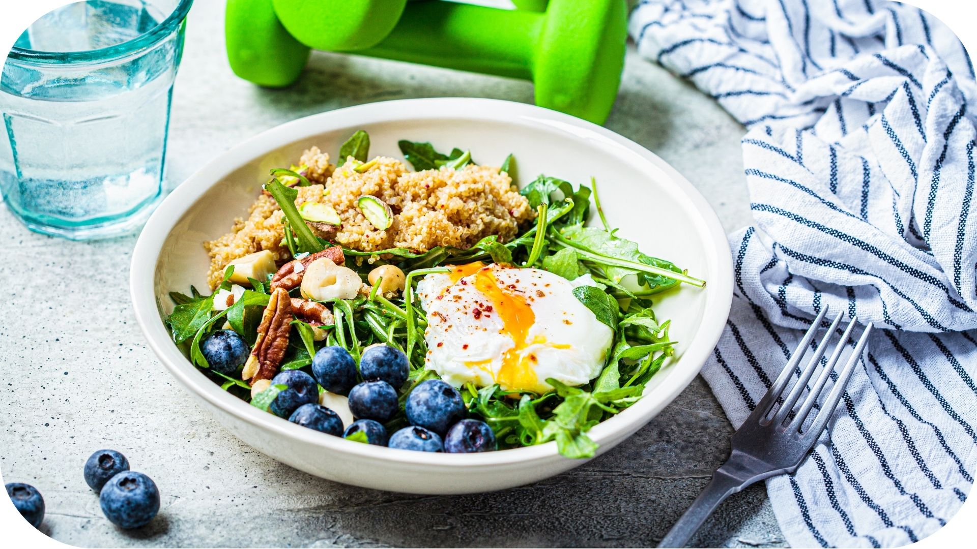 Bowl of healthy salad featuring quinoa, arugula, a poached egg, blueberries, and walnuts, accompanied by water and a green dumbbell