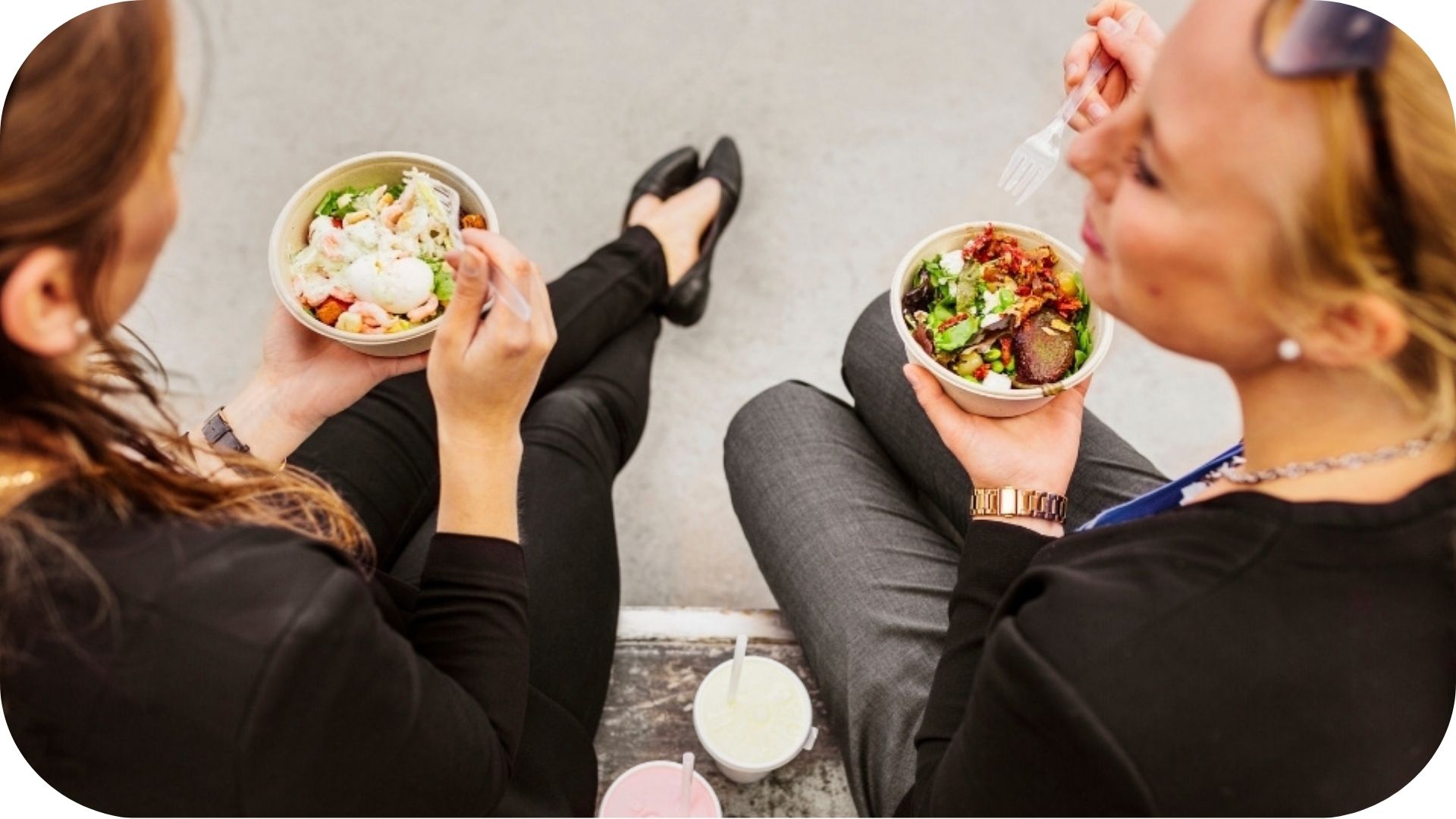 Two women enjoying salads outdoors, each holding a fork and chatting