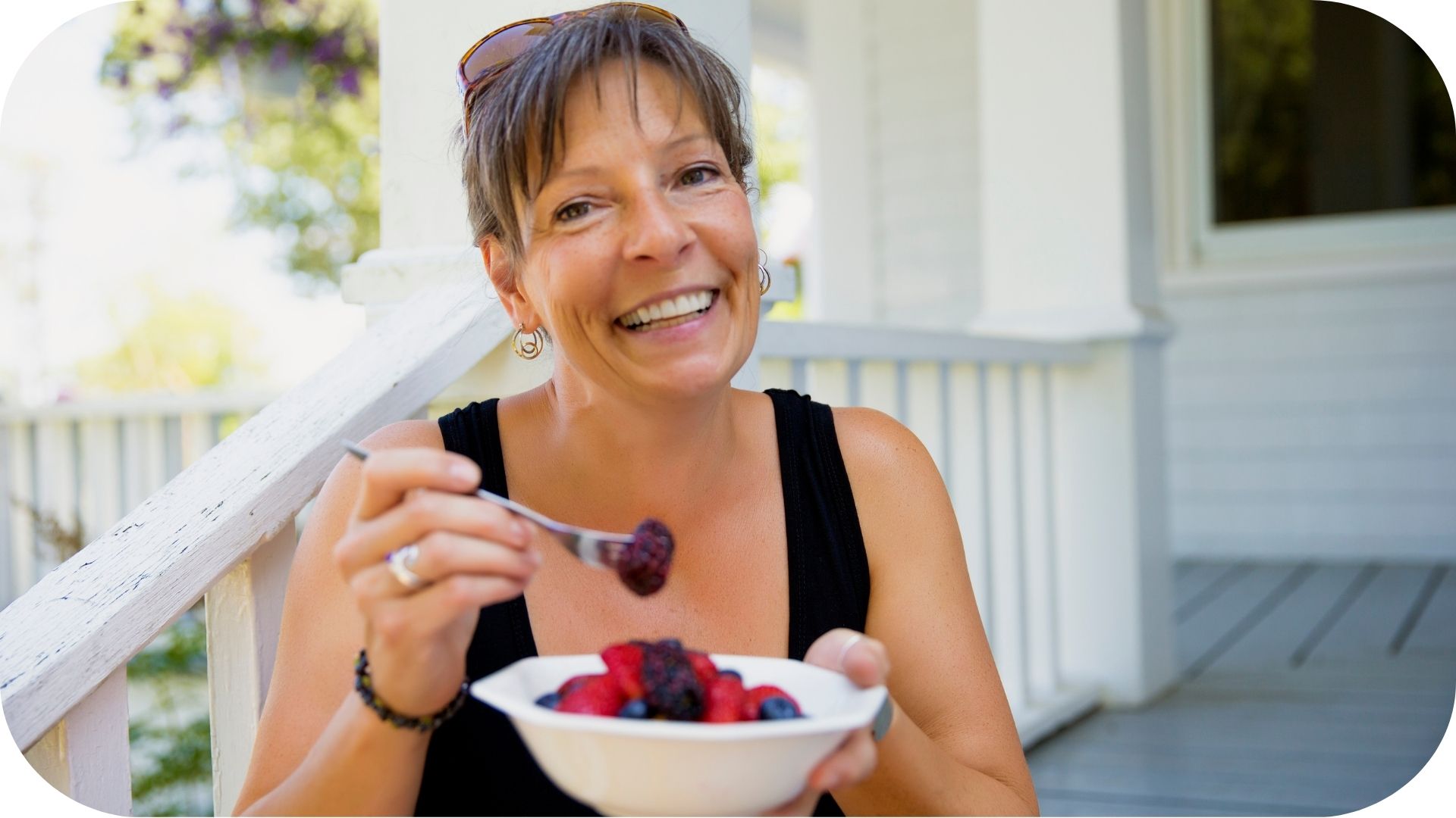 Smiling woman eating a bowl of fresh berries while sitting on a sunny porch