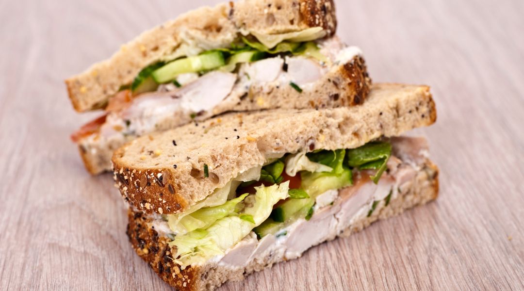 Two halves of a chicken sandwich with whole grain bread, lettuce, cucumber, and a creamy spread, stacked on a wooden surface.