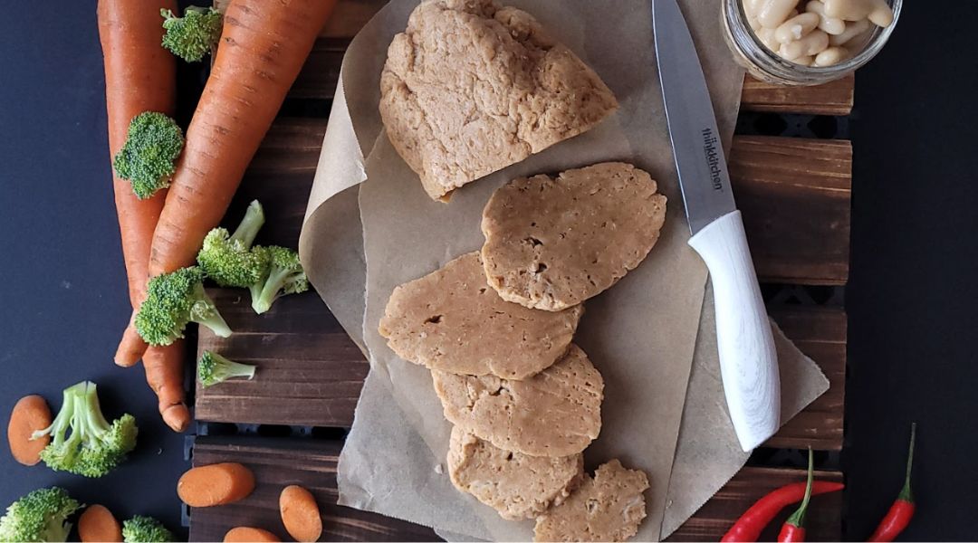 Sliced vegan meat on parchment paper, surrounded by fresh carrots, broccoli, almonds, and a knife on a dark wooden surface.