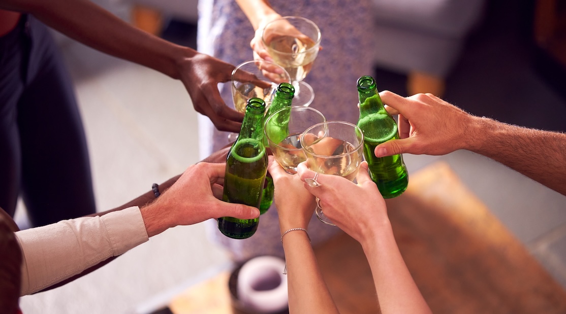 Un groupe de mains diverses trinquant avec des bouteilles de bière verte et des verres de vin dans une ambiance festive. A group of diverse hands toasting with green beer bottles and wine glasses in a celebratory setting.