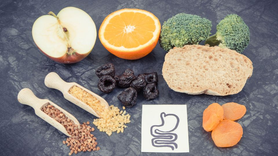 Fibre-rich foods including fruits like apples and oranges, vegetables like broccoli, whole grain bread, dried apricots, prunes, as well as cereals such as buckwheat and brown rice, placed next to an illustration of a stomach to represent potential discomforts related to the presence of FODMAPs.