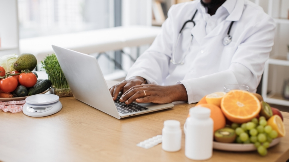 A nutritionist and Registered Dietitian in a white coat works on a laptop with fresh fruits, vegetables, and supplements on the table.