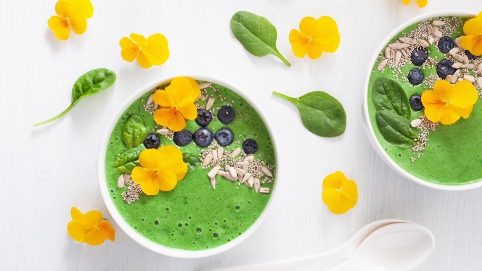 A vibrant green smoothie bowl topped with yellow edible flowers, fresh blueberries, chia seeds, sunflower seeds, and baby spinach leaves. The bowl is arranged on a light background with additional spinach leaves and yellow flowers scattered around for decoration.