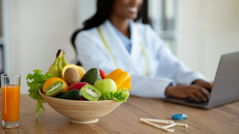 A bowl of fresh fruits, including kiwi, apple, and banana, with a glass of juice on a table, while a nutritionist works on a laptop in the background.