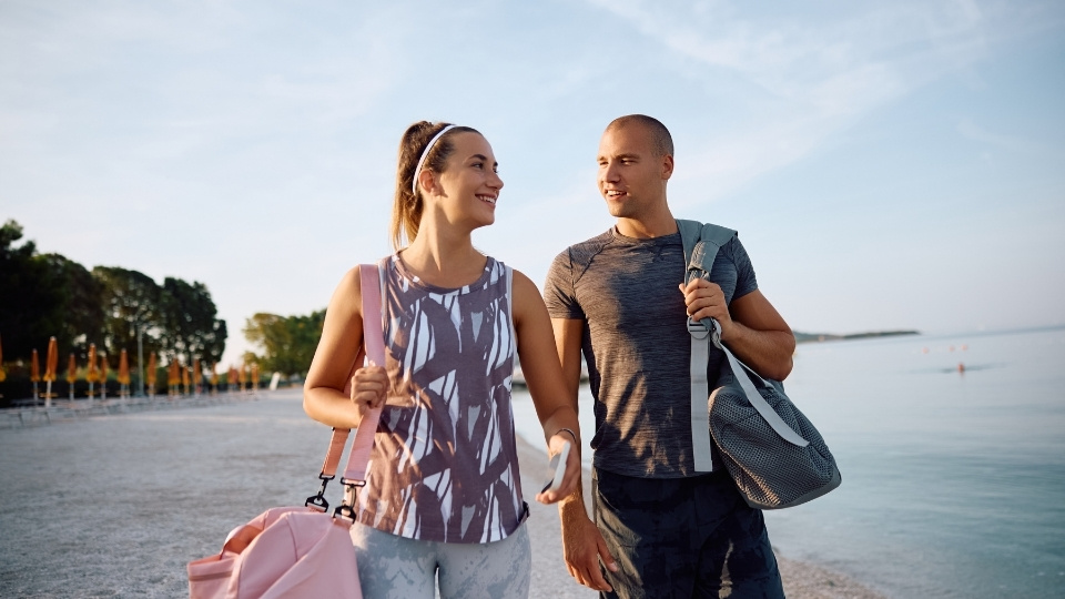 A smiling couple in activewear walks on a beach, each carrying a gym bag, with the sea and trees in the background.