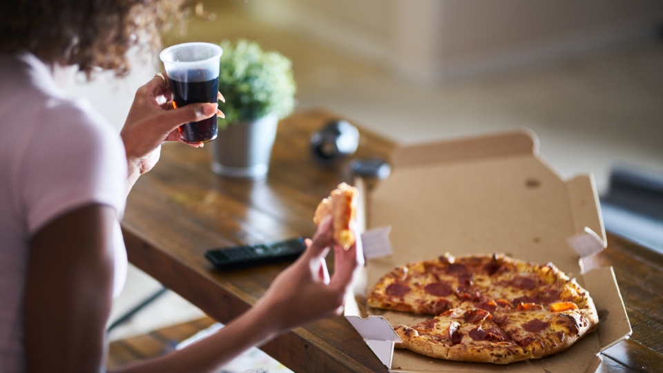 Person holding a slice of pepperoni pizza and a cup of soda, with a pizza box on a wooden table.