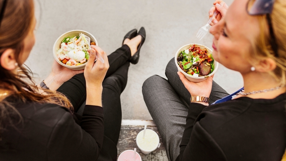 Two women seated outdoors, eating fresh salad bowls with mixed greens and toppings, accompanied by drinks.