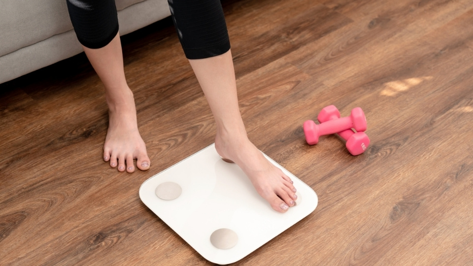 Person stepping on a white scale in a living room with pink dumbbells on the wooden floor nearby.