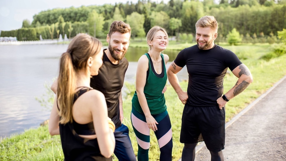 A group of four friends in activewear smiling and chatting outdoors near a lake, with a forested area in the background.