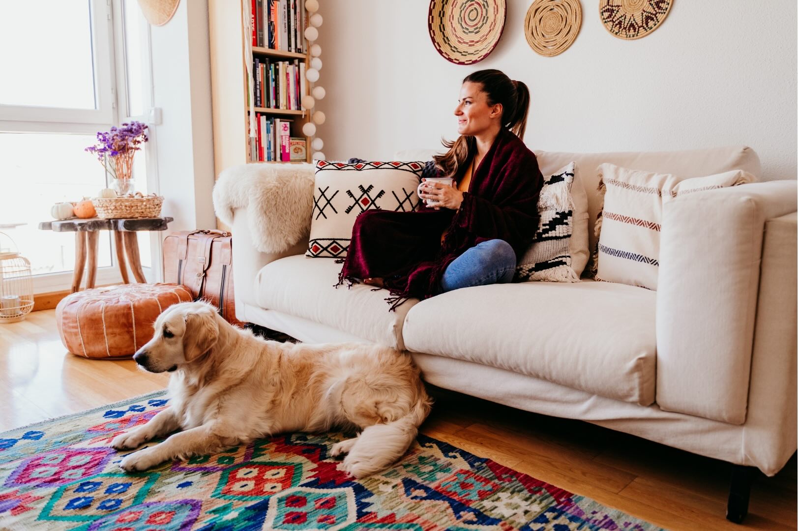 Femme assise sur un canapé avec une tasse, un chien couché sur un tapis coloré devant elle - Woman sitting on a couch with a cup, a dog lying on a colorful rug in front of her