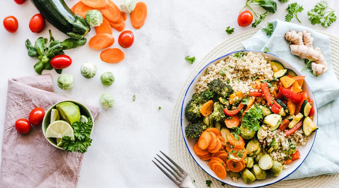 Colorful chopped vegetables scattered on a light background, with a bowl of couscous topped with broccoli, zucchini, carrots, tomatoes, chickpeas, and herbs.