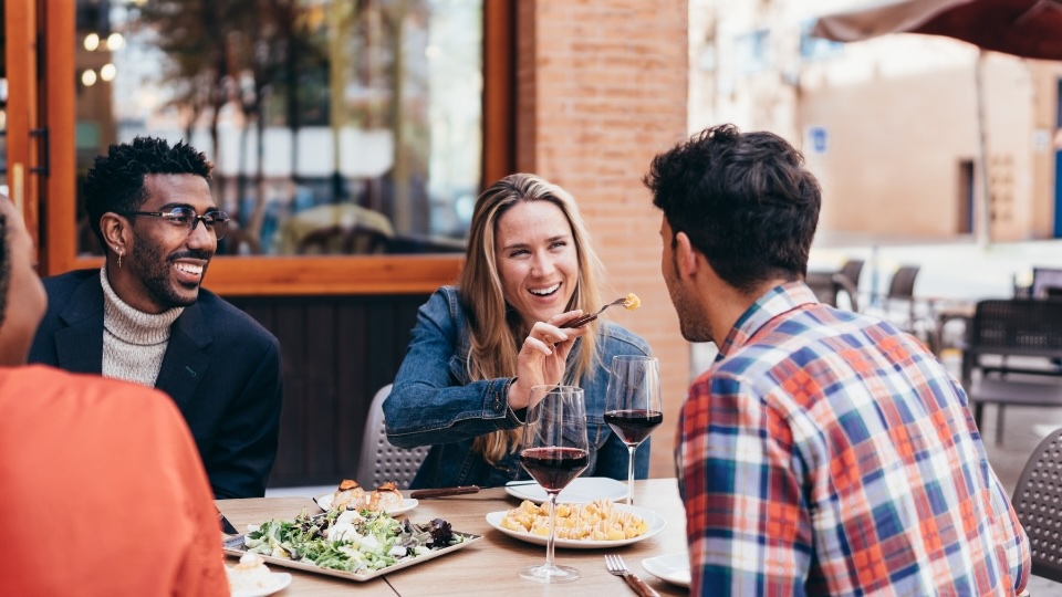 Group of friends enjoying a meal together at an outdoor restaurant, sharing laughs and conversation. The setting reflects a casual and joyful dining experience, highlighting the social benefits of eating out while maintaining healthy choices.