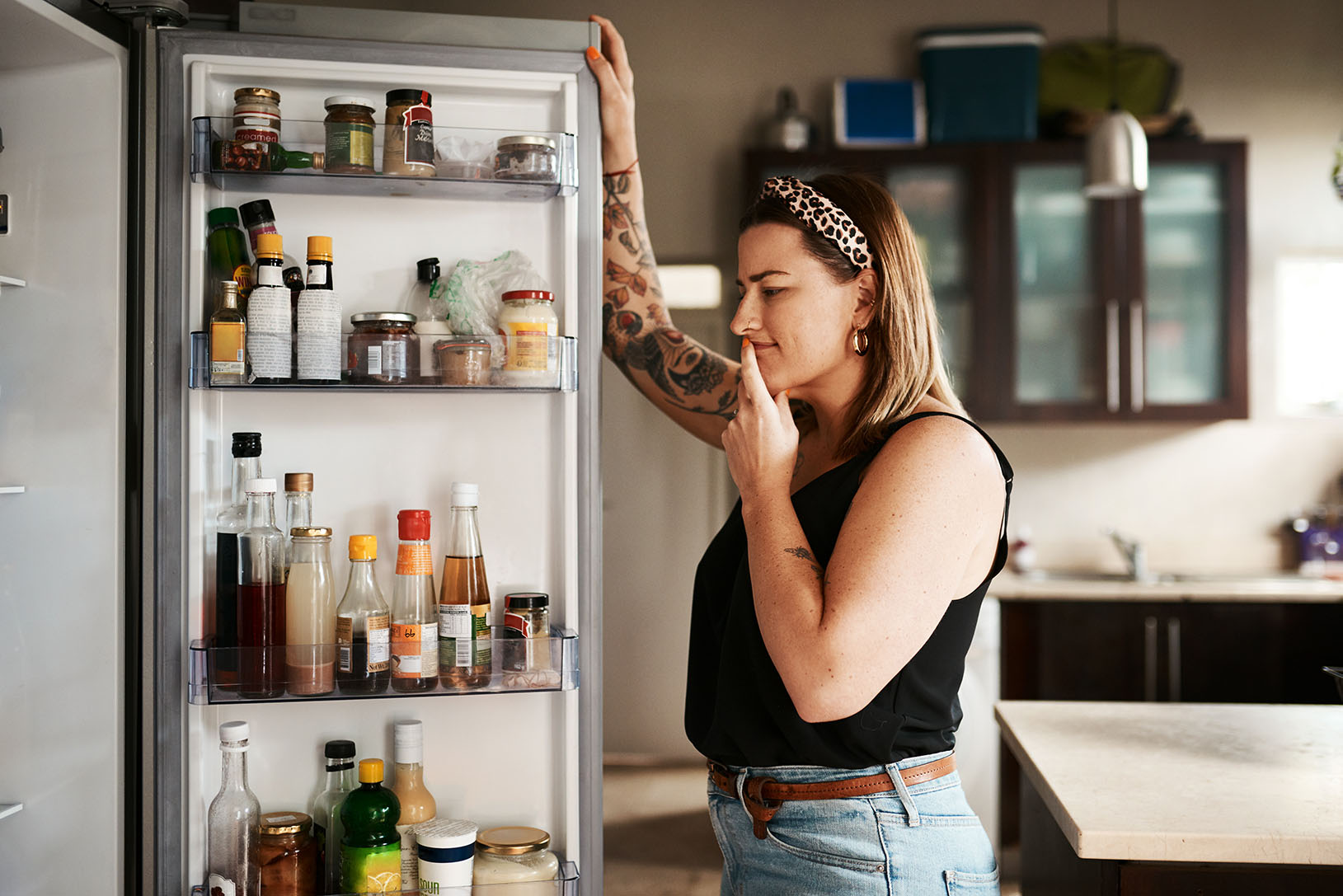 Femme debout devant un réfrigérateur ouvert, réfléchissant en regardant différents aliments et boissons à l'intérieur.  Woman standing in front of an open fridge, thoughtfully looking inside at various food items and drinks.