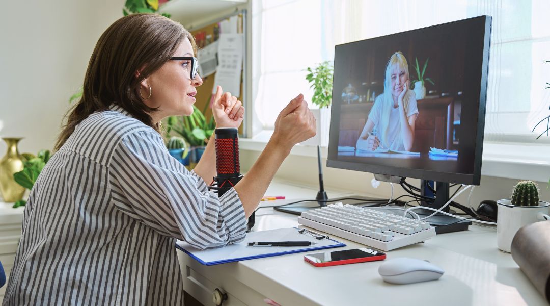 A woman in glasses gestures during a video call with another person on her computer screen, in a home office setting