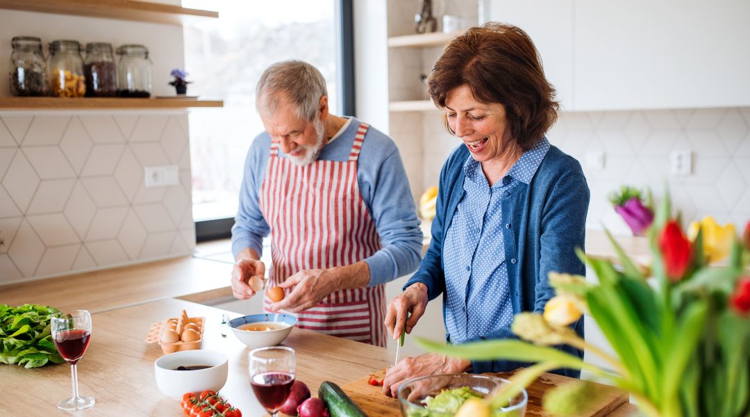 Older couple smiling while preparing vegetables and eggs in a bright kitchen with tulips on the counter