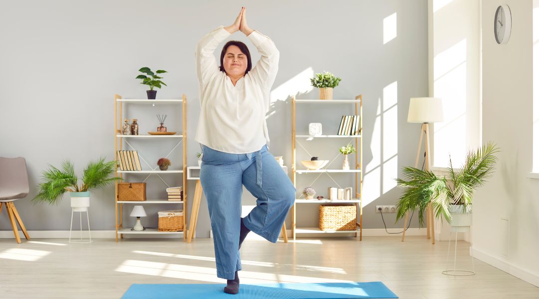 A woman practices yoga in a bright living room, balancing in the tree pose on a blue mat