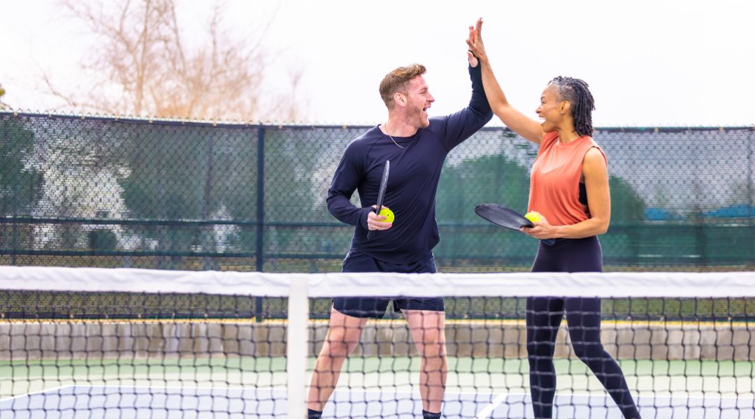 Two people high-fiving on a pickleball court, holding paddles and balls, celebrating a game