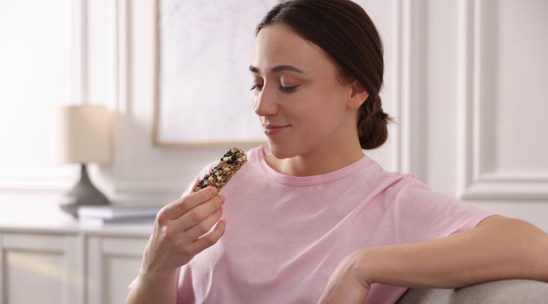 Woman in a pink shirt sitting on a couch, smiling while holding a granola bar