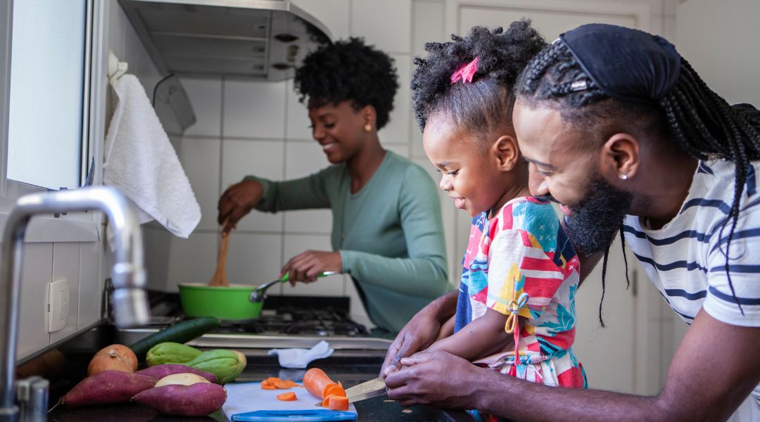 A family of three cooks together in a kitchen, with the father and daughter chopping vegetables and the mother stirring a pot